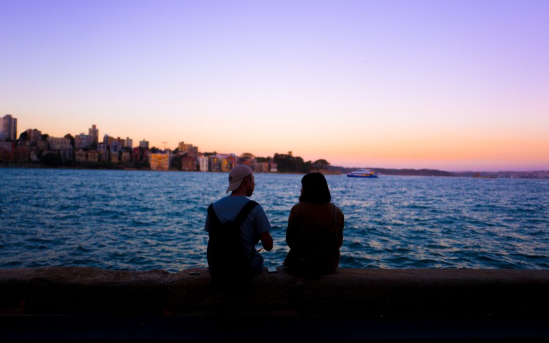 two people chat next to each other sitting across from an ocean with a city line in the distance