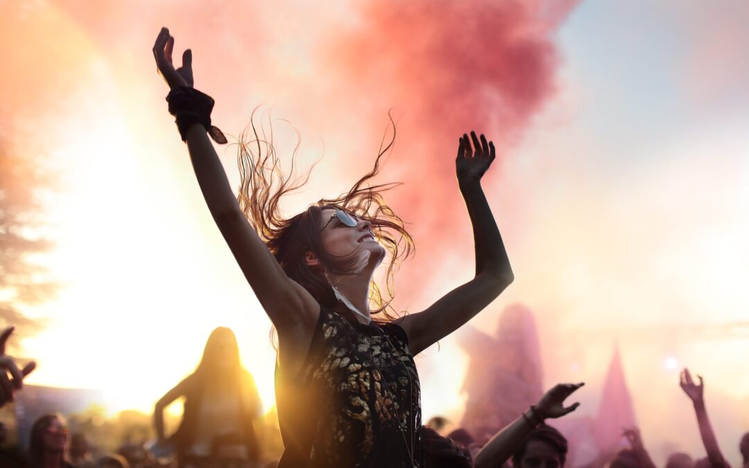 A woman at a concert raises her hands, smoke effects are behind her and its sunset