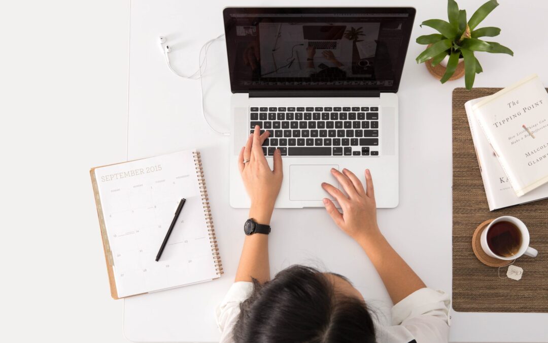 A person sitting at a desk with a notebook, cup of tea, a plant, and a computer