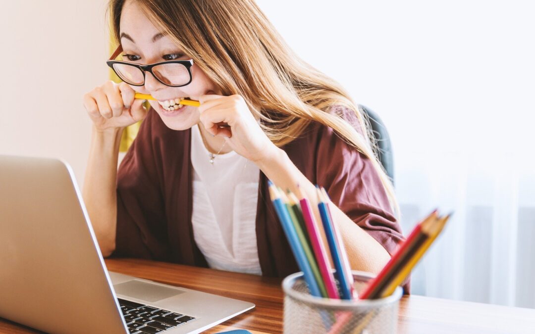 A woman wearing glasses sitting in front of a computer holding a pencil with her teeth