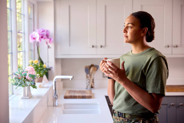 Female American Soldier In Uniform In Kitchen On Home Leave Looking Out Of Window Drinking Coffee