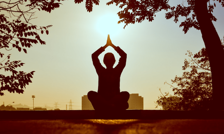 A man doing yoga is clapping his hands while the sun is just above him. He is in a park.