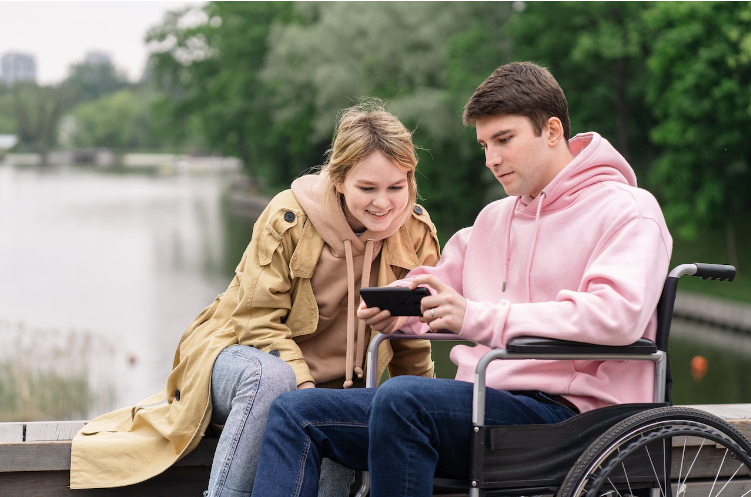 a person in a wheelchair wearing pink is looking at a cel phone while a woman looks on while sitting next to him.