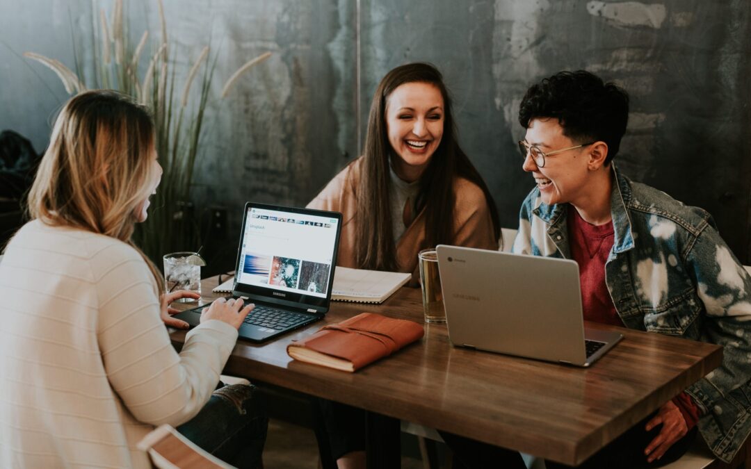 Three people are laughing together at a cafe with laptops open.