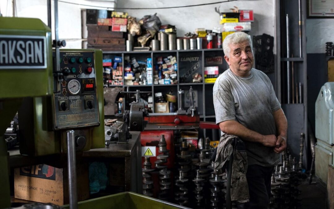 A older man with white hair is looking at the camera while taking a break in a dirty auto mechanic garage.