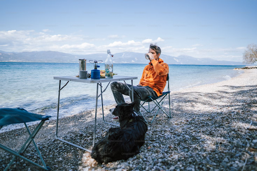 A man sits on the shore of a lake with mountains on the backdrop. He is near his table with drinks and his dog is sitting next to him.